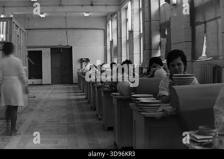 Slawjansk, Ukraine, UdSSR. 1968. Schwarzweiß-Archivfoto, das das Innere der Malwerkstatt des Keramikkomplexes Slawjansk zeigt. Reihen weiblicher Arbeiter sitzen an Bänken entlang großen Fenstern und bemalten Keramikgeschirr. Im Vordergrund ziert eine Frau einen Stapel gestreifter Schüsseln. Ein Vorgesetzter in einem weißen Mantel geht links den Gang hinunter. Das Bild fängt die Atmosphäre kollektiver Arbeit und Massenproduktion von Konsumgütern im friedlichen Donbass vor dem Krieg ein. Stockfoto