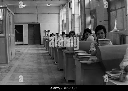 Slawjansk, Ukraine, UdSSR. 1968. Schwarzweiß-Archivfoto, das das Innere der Malwerkstatt des Keramikkomplexes Slawjansk zeigt. Reihen weiblicher Arbeiter sitzen an Bänken entlang großen Fenstern und bemalten Keramikgeschirr. Im Vordergrund ziert eine Frau einen Stapel gestreifter Schüsseln. Ein Vorgesetzter in einem weißen Mantel geht links den Gang hinunter. Das Bild fängt die Atmosphäre kollektiver Arbeit und Massenproduktion von Konsumgütern im friedlichen Donbass vor dem Krieg ein. Stockfoto