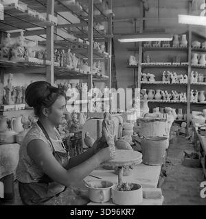 Slawjansk, Ukraine, UdSSR. 1968. Schwarzweiß-Archivfoto, das eine Modelliererin des Keramikkomplexes Slavjansk zeigt, wie sie eine Tonfigur von Hand modelliert. Der Arbeiter, der ein ärmelloses Kleid und eine stilvolle Bienenstockfrisur aus den 1960er Jahren trägt, sitzt an einem Tisch mit Schüsseln aus Ton. Holzregale im Hintergrund sind mit trockenem rohen Keramikstatuen, darunter Vögel und menschliche Figuren, gefüllt. Dieses industrielle Image fängt die Herstellung von Porzellan-Konsumgütern im friedlichen Donbass vor dem Krieg ein. Stockfoto