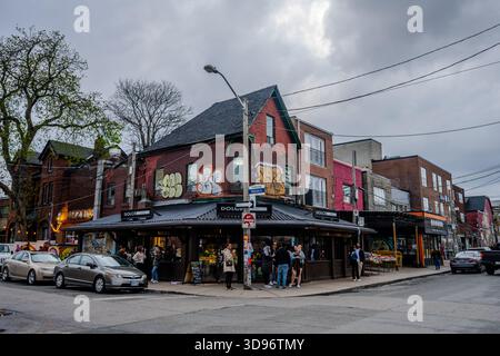 Fußgänger versammeln sich vor Cafés in einem mit Graffiti überdachten Eckgebäude in Toronto, während Autos unter bewölktem Himmel die Stadtstraße säumen. Toronto, Ontario, Stockfoto