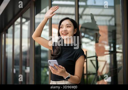 Asiatische Studentin oder Büroangestellte, die vor einem Café, einem Café oder einem Geschäft ein Telefon hält und mit der Hand winkt. Einen Freund treffen, das tägliche Leben, raus gehen. Stockfoto