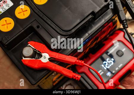 Nahaufnahme einer Kfz-Batterie, die an ein tragbares Ladegerät angeschlossen ist, mit einer roten Klemme am Terminal und der digitalen Spannungsanzeige des Geräts. Stockfoto