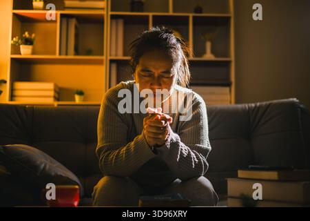 Christlich-religiöse Frau betet nachts auf der Couch im dunklen Wohnzimmer zu Hause. Menschliche Person händet Anbetung, Konzept des Gebets zu Gott. Weiblich Stockfoto