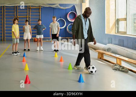 Ein schwarzer Mann demonstrierte vor einer Gruppe von vier Kindern, die in der Schlange stehen, und beobachtete aufmerksam im Fitnessstudio mit bunten Kegeln auf dem Boden Stockfoto