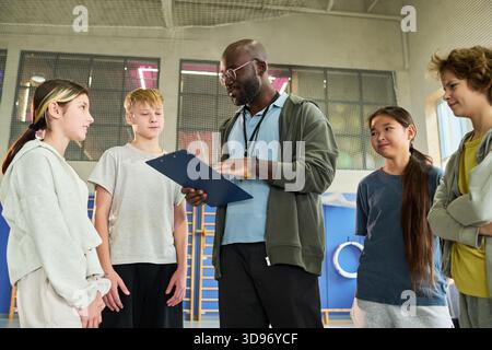 Schwarzer Mann, der verschiedene Gruppen von Teenagern in der Turnhalle unterrichtet, Clipboard hält und Schüler in Diskussionen einbindet, Teenager hören aufmerksam während der sportlichen Aktivität zu Stockfoto