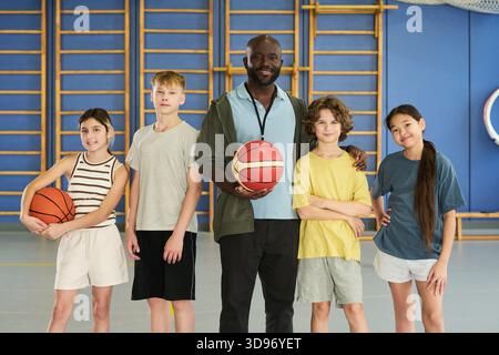 Ein schwarzer Mann steht mit vier Teenagern im Fitnessstudio, die Basketbälle halten, die Gruppe lächelt und vor der Kamera posiert, zwei kaukasische Jungen und zwei Mädchen verschiedener Ethnien stehen zusammen Stockfoto