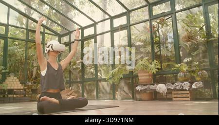 Üben Sie eine Frau, die auf dunkler Yogamatte im Gewächshaus meditiert, mit weißem VR-Headset und Tanktop Stockfoto