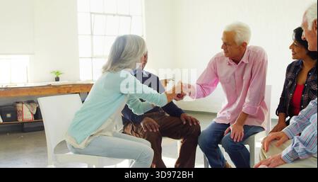 Hände schütteln grauhaarige Frau und weißhaariger Mann im Gemeinschaftsraum, mit weißen Stühlen Stockfoto