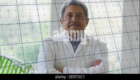 Stehender Senior Mann mit weißem Laborkittel, überkreuzten Armen, Kamera in Klinik, Gitterglas Stockfoto