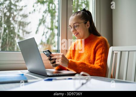 Arbeiten Sie von zu Hause aus mit Telefon und Laptop. Frau mit Computer auf Küchentisch. Unternehmer erstellen geschäftliche Inhalte mit Smartphone. Glücklicher Freelancer. Stockfoto