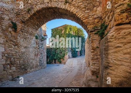 Blick durch einen breiten, alten Steinbogen, der eine sonnenbeschienene, nach oben geneigte Kopfsteinpflasterstraße und mittelalterliche Steinhäuser in Peratallada, Spanien, umrahmt Stockfoto