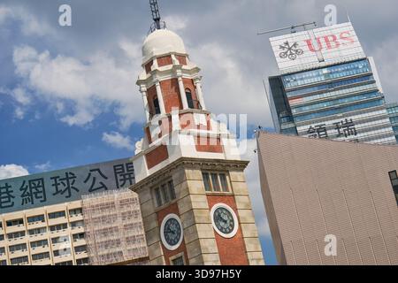 Das historische Wahrzeichen des ehemaligen Kowloon-Canton Railway Clock Tower mit roten Ziegeln und weißer Kuppel, eingerahmt von modernen Wolkenkratzern unter einem hellblauen Himmel. Stockfoto