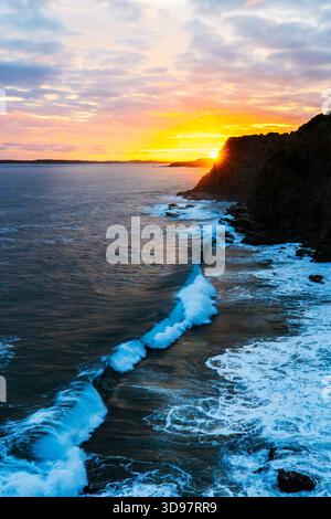 Die kräftigen blauen Wellen des Ozeans prallen bei einem farbenfrohen Sonnenuntergang gegen eine dunkle, zerklüftete Klippe und erleuchten die dramatische Küstenlandschaft. Stockfoto