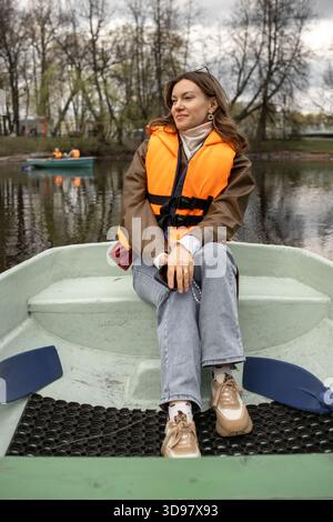 Eine junge Frau mit einer Orange Life Jacket genießt im Frühjahr eine ruhige Bootsfahrt auf einem ruhigen See, der von Bäumen umgeben ist Stockfoto