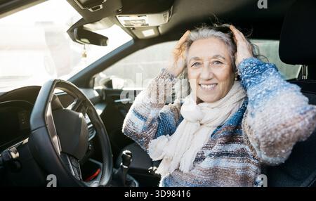 Eine Seniorin sitzt im Auto, lächelt und passt ihr graues Haar an, bereit für eine Reise Stockfoto