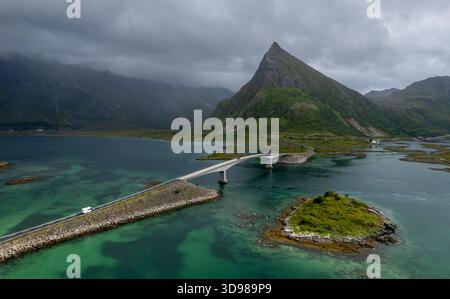 Ein steiler Berggipfel erhebt sich dramatisch aus einem Küstengebiet, umgeben von grünen Hügeln und einer Brücke, die das Wasser überquert. Bedeckter, stimmungsvoller Himmel. Flakstad br Stockfoto