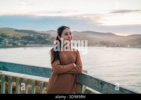Schöne asiatische Frau in braunem Pullover, die am pazifischen Meer mit Bergblick am Abend in Wellington, Neuseeland, steht Stockfoto