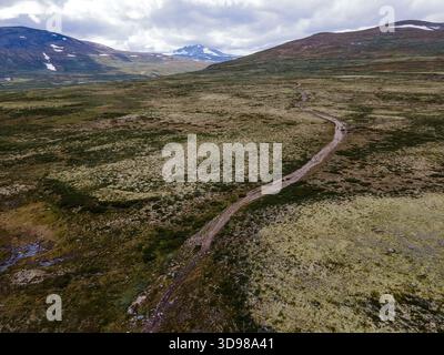 Aus der Vogelperspektive eines gewundenen Pfades, der durch die vielfältigen Strukturen von Rondanes Landschaft führt, im Kontrast zu den weit entfernten, schneebedeckten Berggipfeln, Do Stockfoto