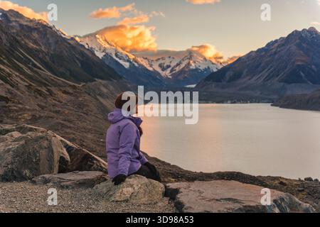 Eine Reisende Frau in lila Jacke und Hut sitzt am Abend auf Felsen mit Blick auf den Tasman Lake Gletscher im Mount Cook Nationalpark, Neuseeland Stockfoto