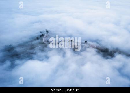 Blick aus der Vogelperspektive auf das Nebelmeer über den Berggipfel, die geschwungene Straße und verstreute Bäume, die eine traumhafte und stimmungsvolle Landschaft schaffen Stockfoto