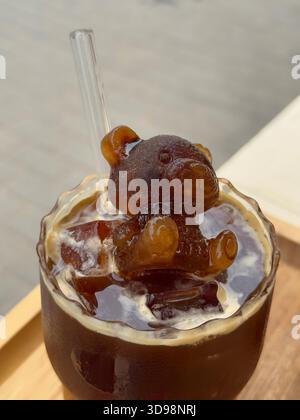 Erfrischender Cold Brew Kaffee serviert in transparentem Glas mit bärenförmigem Eiswürfel und umweltfreundlichem Glasstroh auf hellem Hintergrund Stockfoto