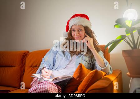 Frau mit weihnachtsmütze, die direkt in die Kamera blickt, während sie Gedanken in ein Notizbuch schreibt, ihre weihnachtswunschliste auf einer gemütlichen Couch erstellt und damit Urlaubsplanung und -Vorfreude symbolisiert Stockfoto
