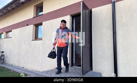 Ein Mann in einer grauen und orangefarbenen Jacke tritt aus einem Gebäude mit einem schwarzen Seesack und schließt die Tür hinter ihm, während er seinen Tag auf einem gepflasterten Spaziergang beginnt Stockfoto