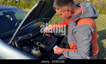 Ein Mann in einer grauen und orangefarbenen Jacke überprüft den Ölstand mit einem Messstab unter der geöffneten Motorhaube eines Autos bei einer Straßenkontrolle in einem grasbewachsenen ländlichen Areal Stockfoto