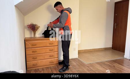 Ein Mann in einer grauen und orangen Jacke steht neben einer hölzernen Kommode, sichert einen schwarzen Seesack mit getrockneten Blumen in einer Vase neben ihm und bereitet sich darauf vor zu gehen Stockfoto