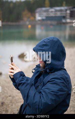 Weibliche Touristenin in blauer Kapuzenjacke, die an einem regnerischen Herbsttag ein Bild von einem malerischen Seeblick mit einem Mobiltelefon macht. Stockfoto