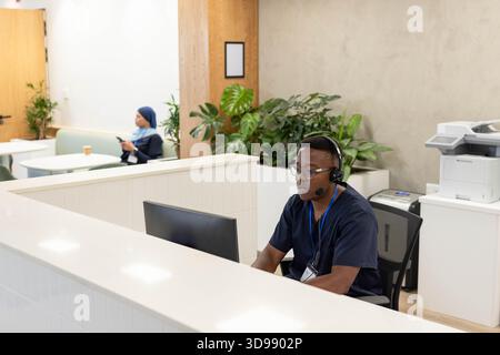 Afroamerikanischer Mann am Schreibtisch im Schrubben mit Headset, das am Computer arbeitet, während eine Frau das Telefon hält Stockfoto