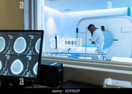 Patient, der auf dem CT-Tisch sitzt und den Kopf hält, Kontrollraummonitor mit Gehirnscans, blaue Gantry Stockfoto