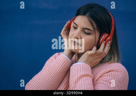 Selbstbewusste junge Frau in rosa Pullover hört Musik mit roten Kopfhörern und entspannt sich an einer hellblauen Wand. Stockfoto