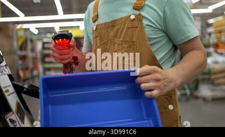 Professionelle Bauarbeiter in braunen Overalls und hellgrünen T-Shirts scannen blaue Farbschalen an der Selbstbedienungskasse in einem Baumarkt mit Barcode-Scanner Stockfoto