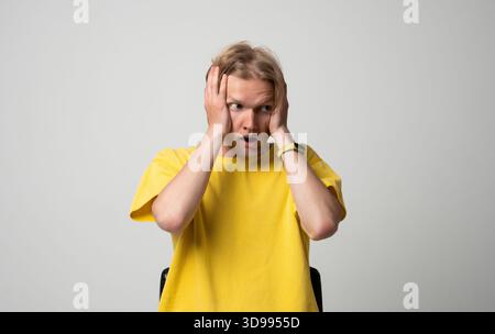 Schockierter junger Mann, der seine Wangen mit beiden Händen hält und mit breiten Augen in neutraler Studioumgebung reagiert Stockfoto