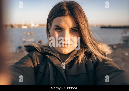 Porträt einer lächelnden jungen Frau, die bei Sonnenuntergang auf einem Küstengang ein Selfie mit ihrem Smartphone macht, warme Jacke und städtischer Hafen im Hinterland Stockfoto