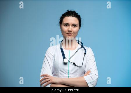 Ärztin mit Stethoskop posiert selbstbewusst vor blauem Hintergrund in Berufsuniform Stockfoto