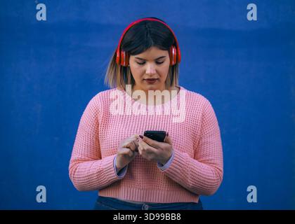 Selbstbewusste junge Frau in rosa Pullover, die Musik hört und ihr Smartphone benutzt, steht draußen vor einem auffälligen blauen Hintergrund. Stockfoto