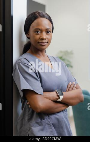 Afroamerikanische Frau, die sich in der Klinik an die Wand lehnt, mit grauen Peelings und Smartwatch in der Nähe eines blaugrünen Stuhls Stockfoto