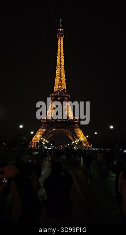 Der Eiffelturm leuchtet nachts in Paris, Frankreich, mit Reflexionen auf nassem Gehweg und lebhafter Menschenmenge. Stockfoto