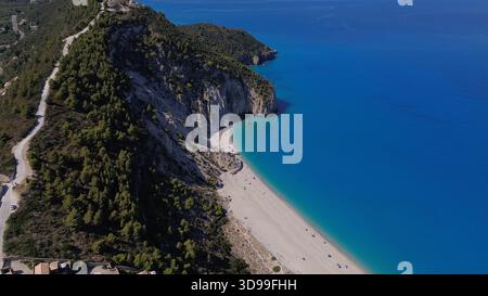 Blick aus der Vogelperspektive auf den abgeschiedenen Sandstrand mit türkisfarbenem Meer und bewaldeter Klippenstraße unter klarem Himmel. Stockfoto