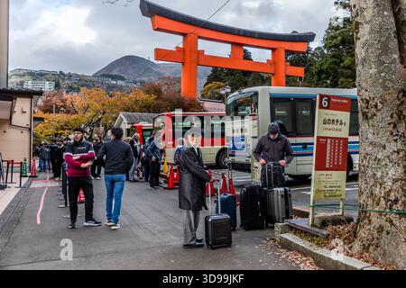 Touristen mit Koffern warten vor einem großen roten Torii-Tor in Hakone auf Shuttlebusse zu den umliegenden Ryokans und Hotels. Das Torii markiert den Eingang zum Hakone-jinja, einem der ältesten Shintō-Schreine der Region, der im Jahr 757 gegründet wurde. Hakone, Präfektur Kanagawa, Japan Stockfoto
