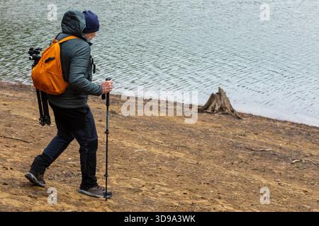Ein Alleinreisender spaziert am Sandstrand eines ruhigen Sees entlang. Ausgestattet mit einem Stativ und einer Kamera, fängt er die ruhige Schönheit ein, während die Dämmerung sich beruhigt Stockfoto