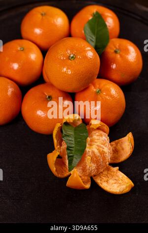 Mandarinen befinden sich auf einer dunklen Oberfläche, eine teilweise geschälte Frucht mit sichtbaren Segmenten und ein frisches grünes Blatt an der Schale, das die Heilung hervorhebt Stockfoto