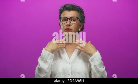 Ältere Frau mit grauem Haar und Brille entspannt sich vor einem leuchtend rosa Hintergrund, wobei sie sanft ihre Hände zur Selbstpflege und zur Linderung von Verspannungen in einem c Stockfoto