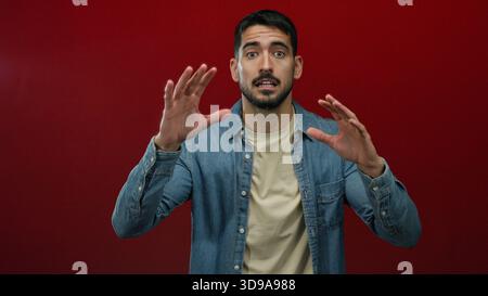 Junger hispanischer Mann in lässiger Kleidung posiert selbstbewusst vor einem leuchtend roten Hintergrund und drückt verschiedene Emotionen mit dynamischen Handgesten aus. Stockfoto