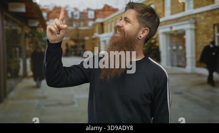 Junger bärtiger Mann mit schwarzem Sweatshirt, der mit dem Zeigefinger nach oben auf die geschäftige Stadtstraße zeigt, gesäumt von Backsteinläden; Hoffnung auf Optimismus. Stockfoto