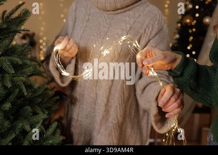 Nahaufnahme von zwei Frauen in gemütlichen Pullover, die leuchtende Lichterketten halten, während sie Tannenbäume in einem warmen, festlichen Innenraum dekorieren. Stockfoto