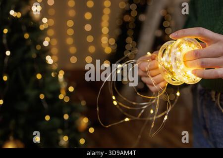 Nahaufnahme einer Frau, die helle Lichter von der Walze mit verschwommenem Weihnachtsbaum und goldenem Bokeh in gemütlichem Zuhause entwirrt. Stockfoto