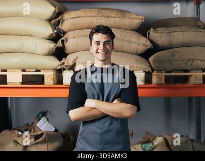 Porträt eines jungen selbstbewussten männlichen Arbeiters auf der Schürze im Lager. Ein Mann mit gekreuzten Armen, der eine Schürze trägt, steht im Lager. Stockfoto